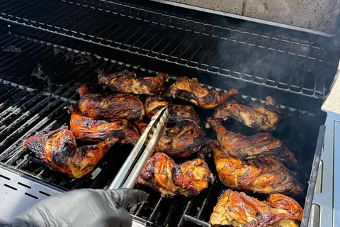 Jerk chicken quarters searing on an Alpha Katering grill, tongs turning a piece