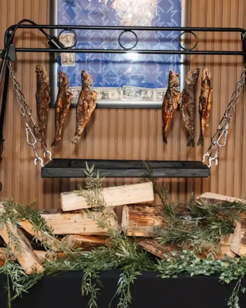 A carved fish display hung from a black frame above firewood and fresh rosemary